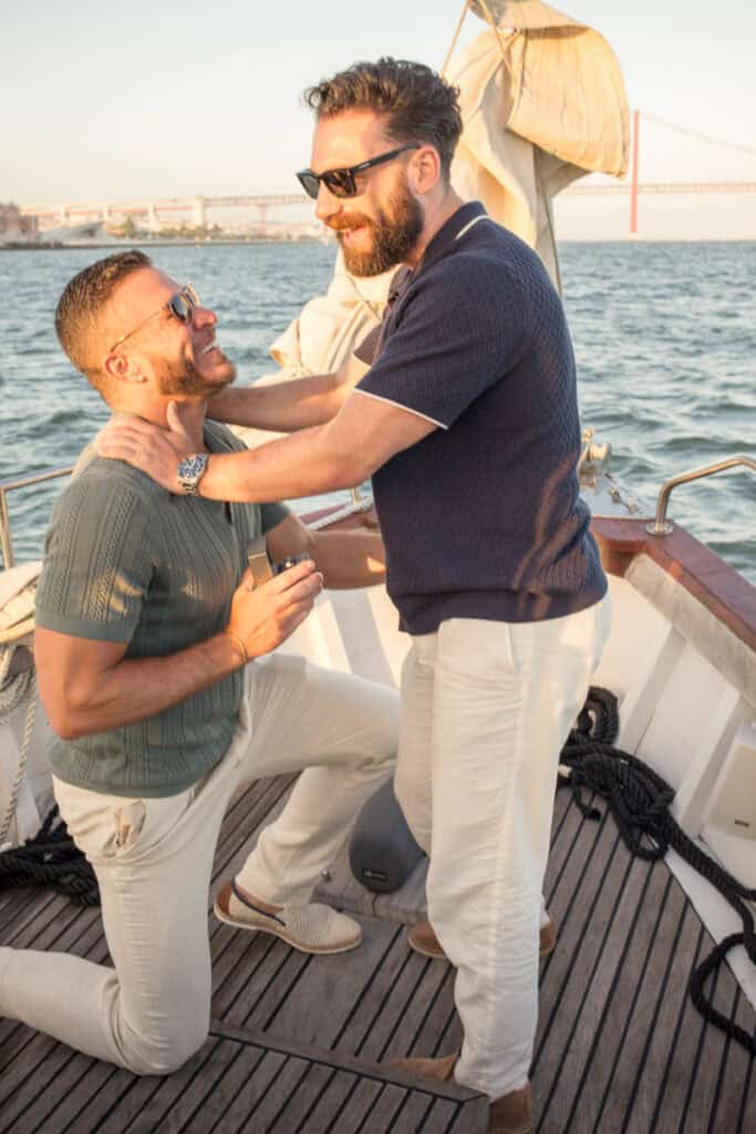 Een romantisch Lissabon huwelijksaanzoek op een zeilboot tijdens zonsondergang, met de iconische 25 april-brug op de achtergrond terwijl de ene man op zijn knie gaat voor de andere.