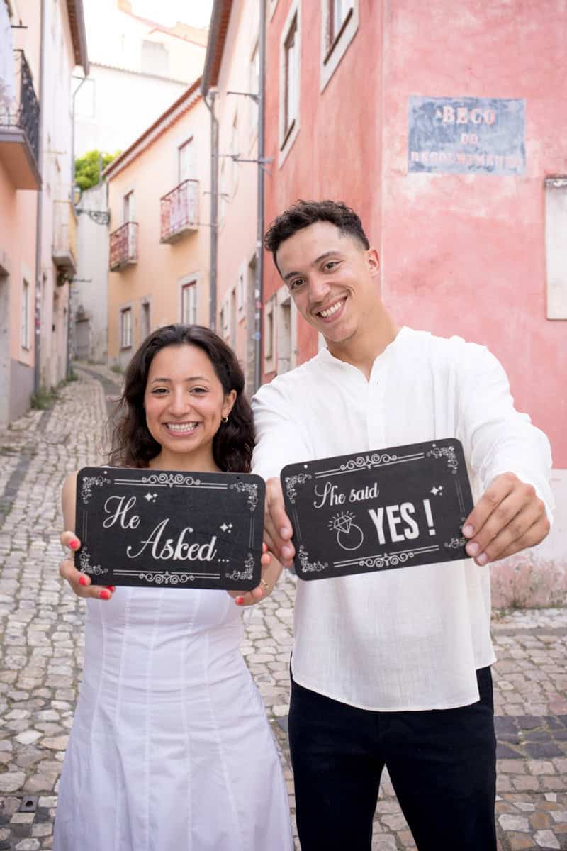 Een stralend stel viert hun huwelijksaanzoek in Lissabon in een kleurrijke steeg van Alfama. Ze houden bordjes vast met de tekst "He Asked..." en "She said YES!".