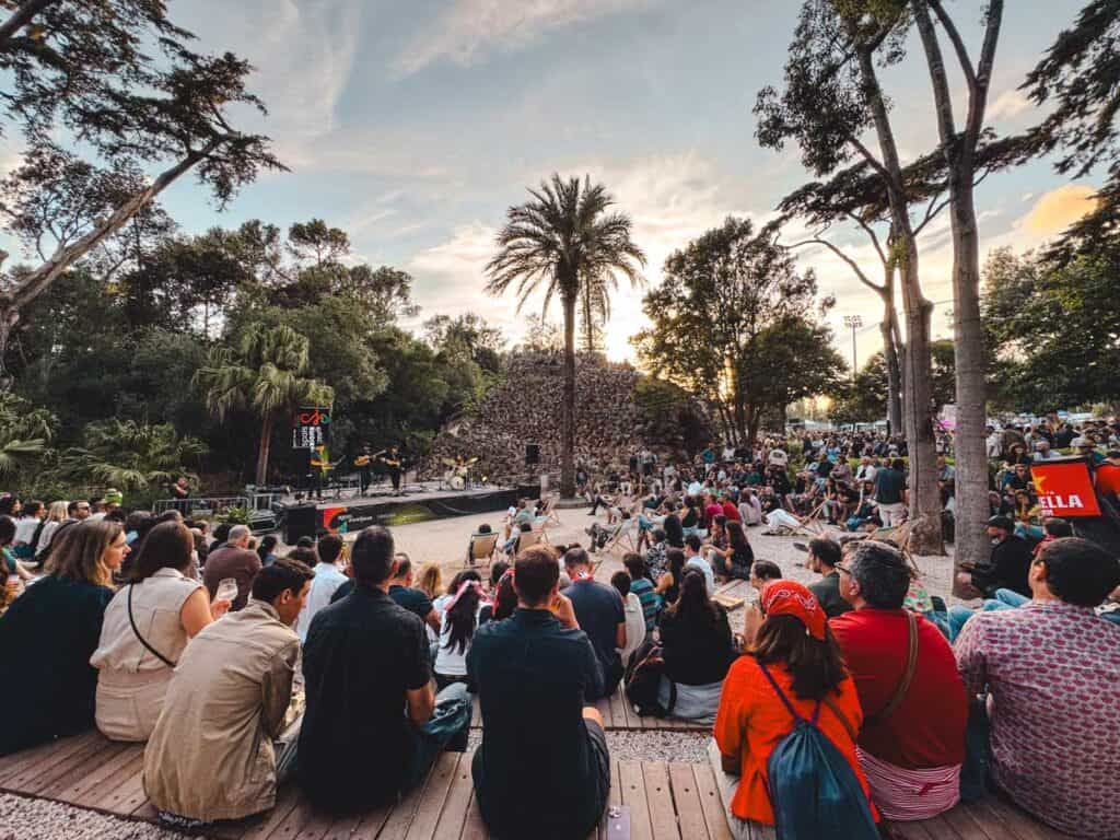 A large crowd watches a live band perform at the Ageas Cooljazz festival in Cascais in a wooded outdoor amphitheatre at sunset. Tall trees and a palm frame the stage beside a rough stone structure while signs read "Ageas Cooljazz" and "Stella".