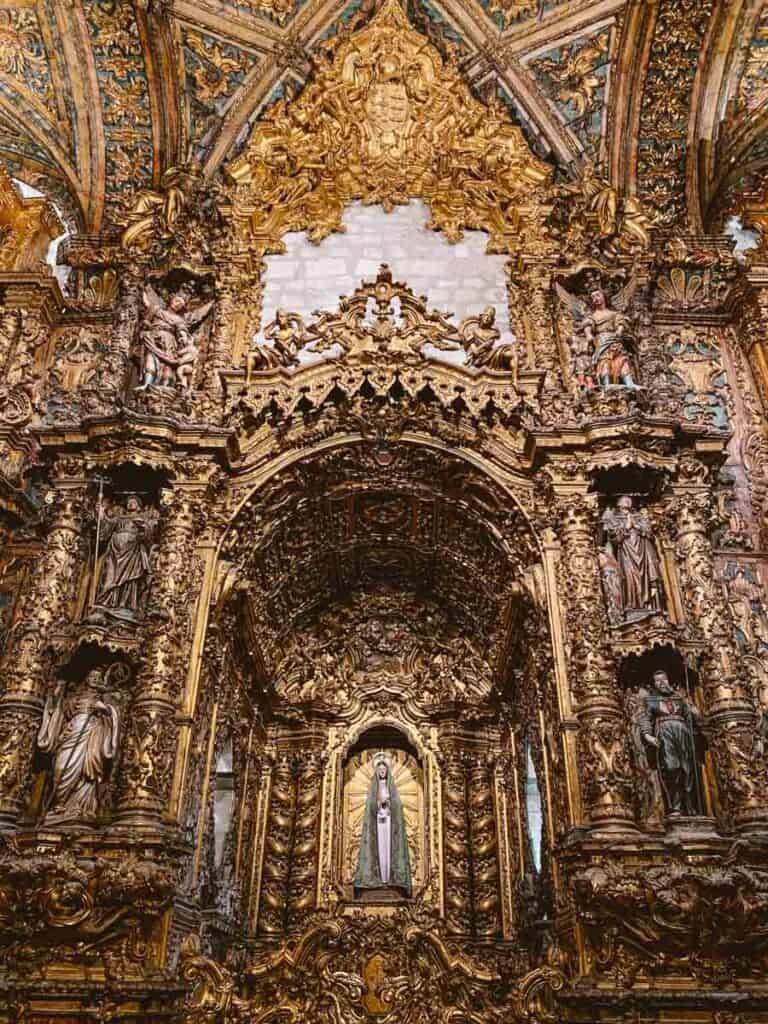 Retábulo de Nossa Senhora da Rosa ricamente decorado em talha dourada com esculturas de santos distribuídas em colunas e nichos ao redor. No centro, a imagem de Nossa Senhora em destaque é envolvida por ornamentos barrocos e um arco detalhado que reforça a grandiosidade e riqueza artística do altar.
