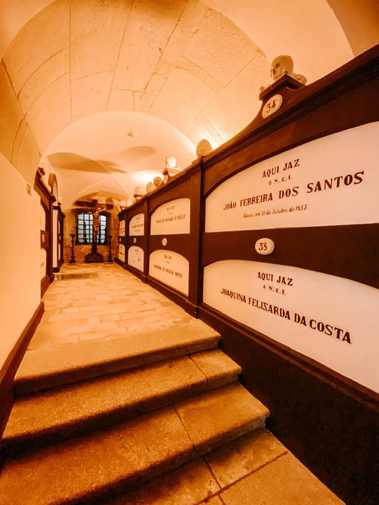 Corredor da cripta da igreja São Francisco Porto com nichos funerários nas paredes identificados com nomes como "AQUI JAZ JOÃO FERREIRA DOS SANTOS" e "AQUI JAZ JOAQUINA FELISARDA DA COSTA". A escadaria de pedra conduz até um crucifixo ao fundo, iluminado por luz quente que reforça a atmosfera solene do espaço.