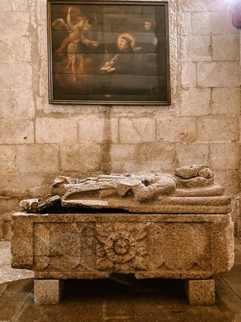 Capela da família Carneiro no Porto com túmulo de pedra esculpido mostrando uma figura deitada sobre a lápide. Ao fundo, uma parede de blocos de pedra sustenta um quadro religioso, criando um ambiente silencioso e histórico dentro da igreja.