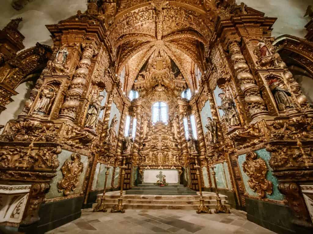 Interior da igreja de São Francisco no Porto com altar-mor totalmente revestido em talha dourada e colunas ornamentadas repletas de esculturas religiosas. A luz entra pelas janelas ao fundo iluminando o espaço, destacando a riqueza barroca e os detalhes exuberantes do altar.