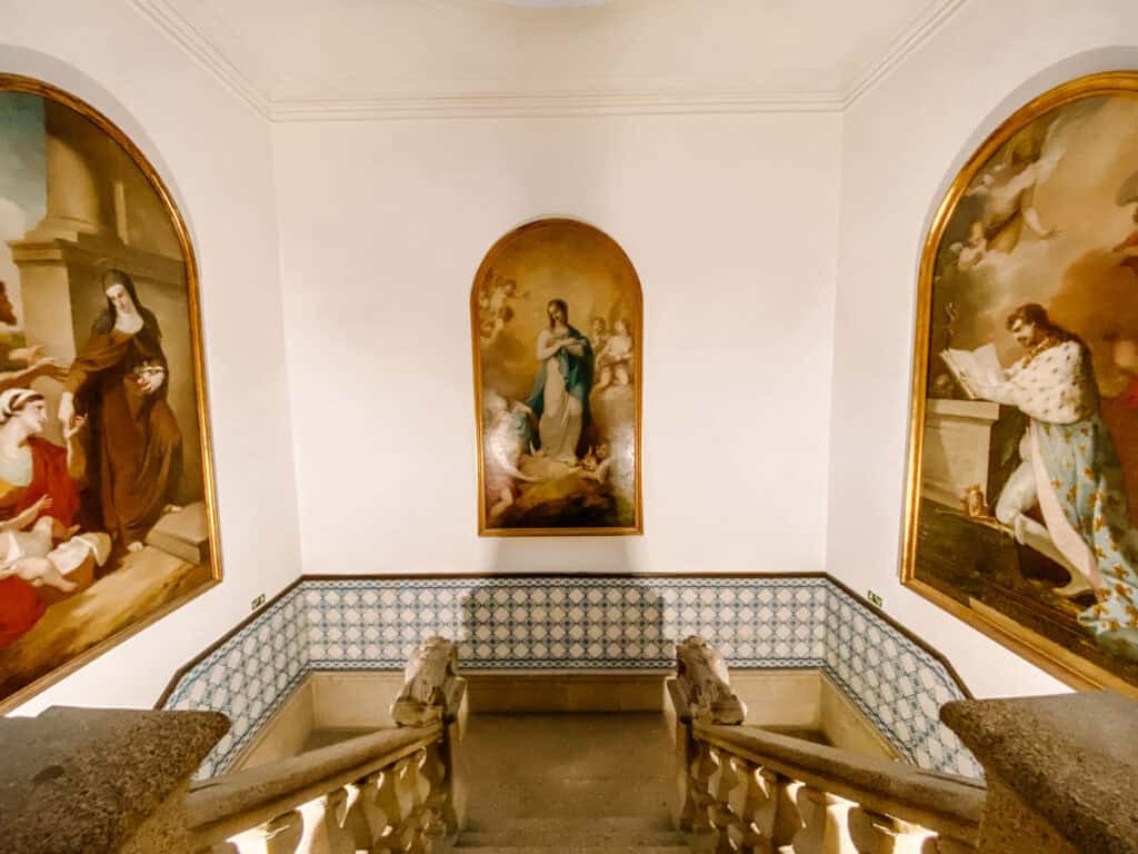 Escadaria interna da igreja de São Francisco Porto com paredes brancas decoradas por azulejos e três grandes pinturas religiosas emolduradas. As telas mostram cenas sacras com figuras centrais em destaque, criando um ambiente calmo que conecta diferentes espaços da igreja.