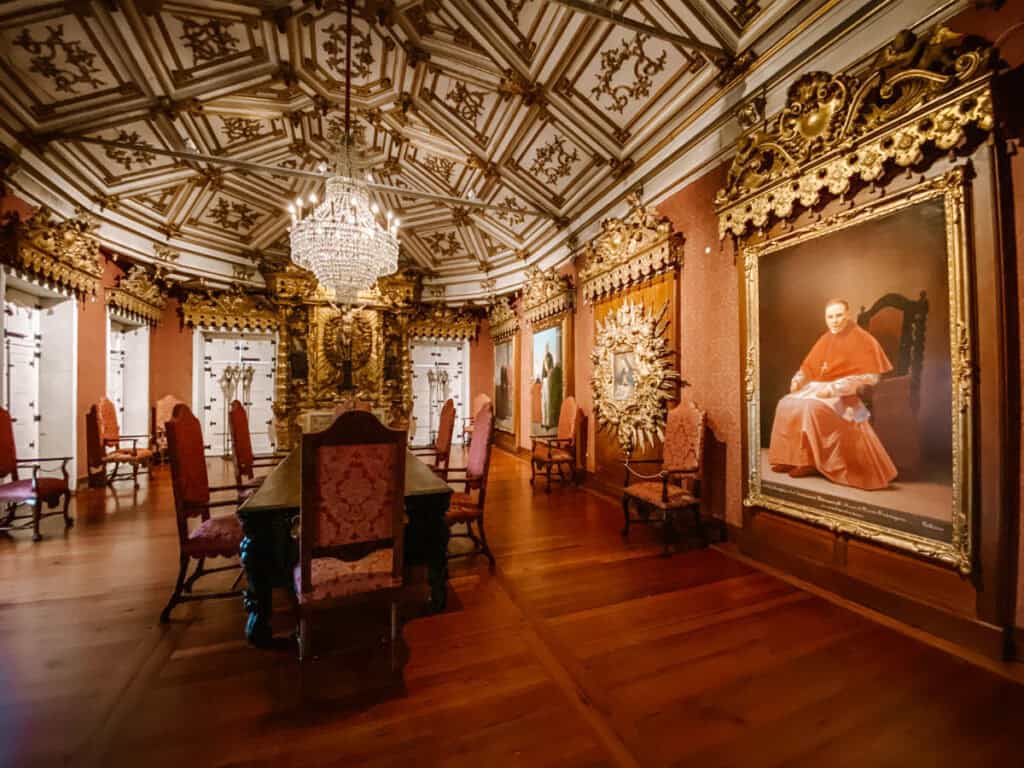 Sala ornamentada da igreja de São Francisco Porto com mesa longa rodeada por cadeiras estofadas e lustre de cristal pendurado no teto decorado. As paredes exibem grandes retratos religiosos em molduras douradas, criando um ambiente histórico e elegante dentro do complexo da igreja.