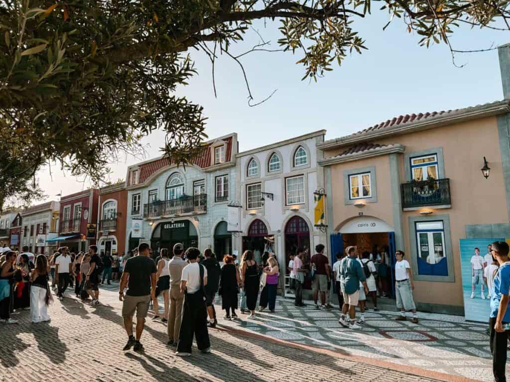 A busy pedestrian street lined with pastel shopfronts and patterned paving fills with visitors in the afternoon sun. Storefront signs read "Gelateria" and "carris" as crowds explore the area during a trip to NOS Alive Lisbon.