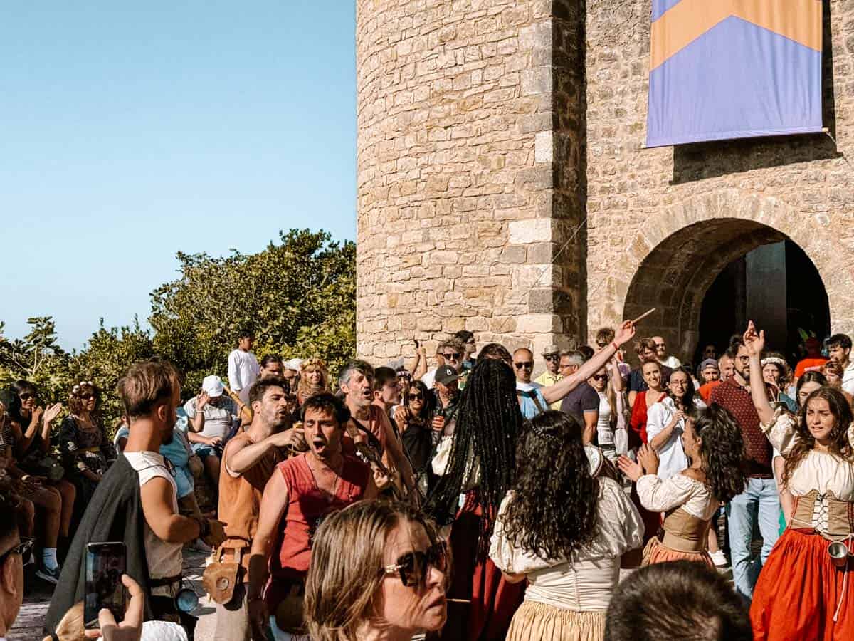 Crowd gathered outside a historic stone building in Portugal dancing and clapping during a lively Portugal festivals celebration. Performers in rustic costumes lead the festivities while onlookers smile, film on phones, and join in under bright sunshine.