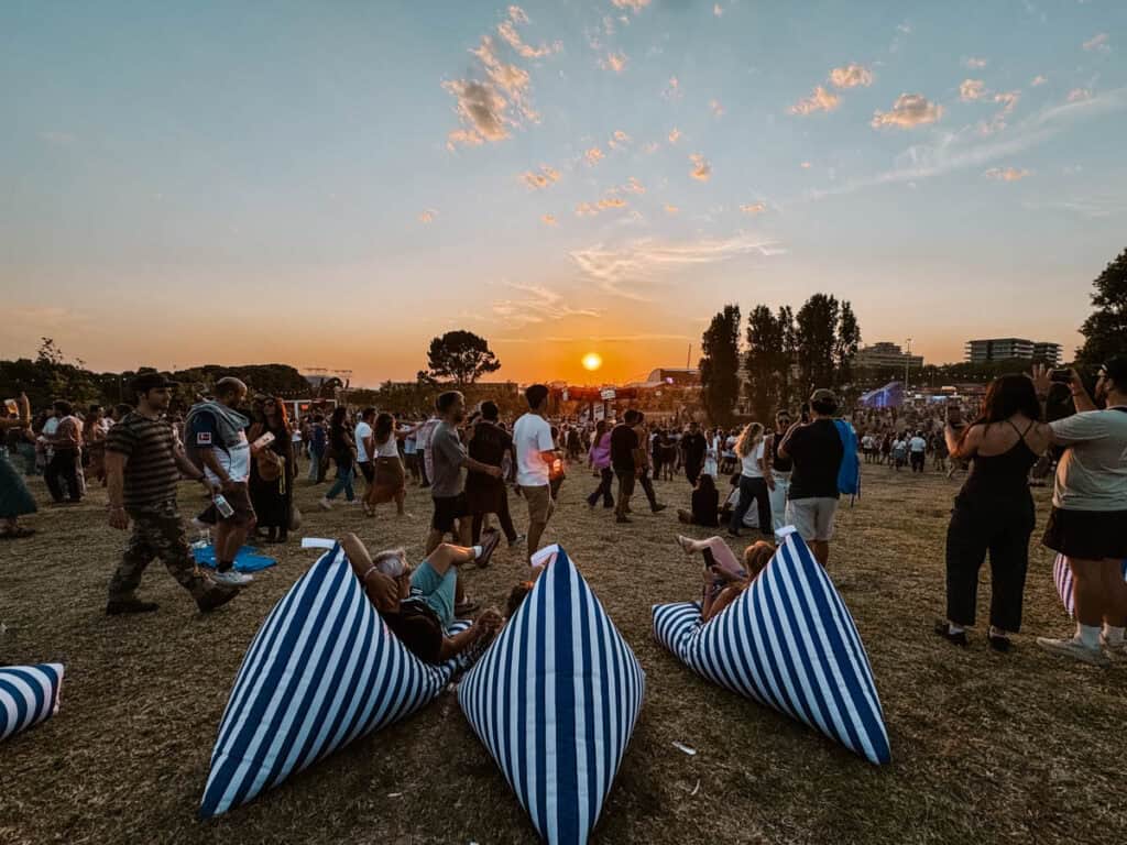 Festivalgoers relax on blue and white striped beanbags while crowds cross a dry grassy field at Primavera Sound Festival Porto. The sun sets behind distant stages and trees under a pale blue sky with pink clouds.