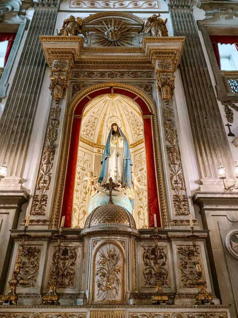 Altar lateral da igreja de São Francisco no Porto com escultura de Nossa Senhora em destaque dentro de um nicho iluminado com fundo dourado e vermelho. A estrutura ao redor apresenta colunas e detalhes esculpidos em dourado, com castiçais e elementos decorativos que reforçam a elegância do espaço religioso.