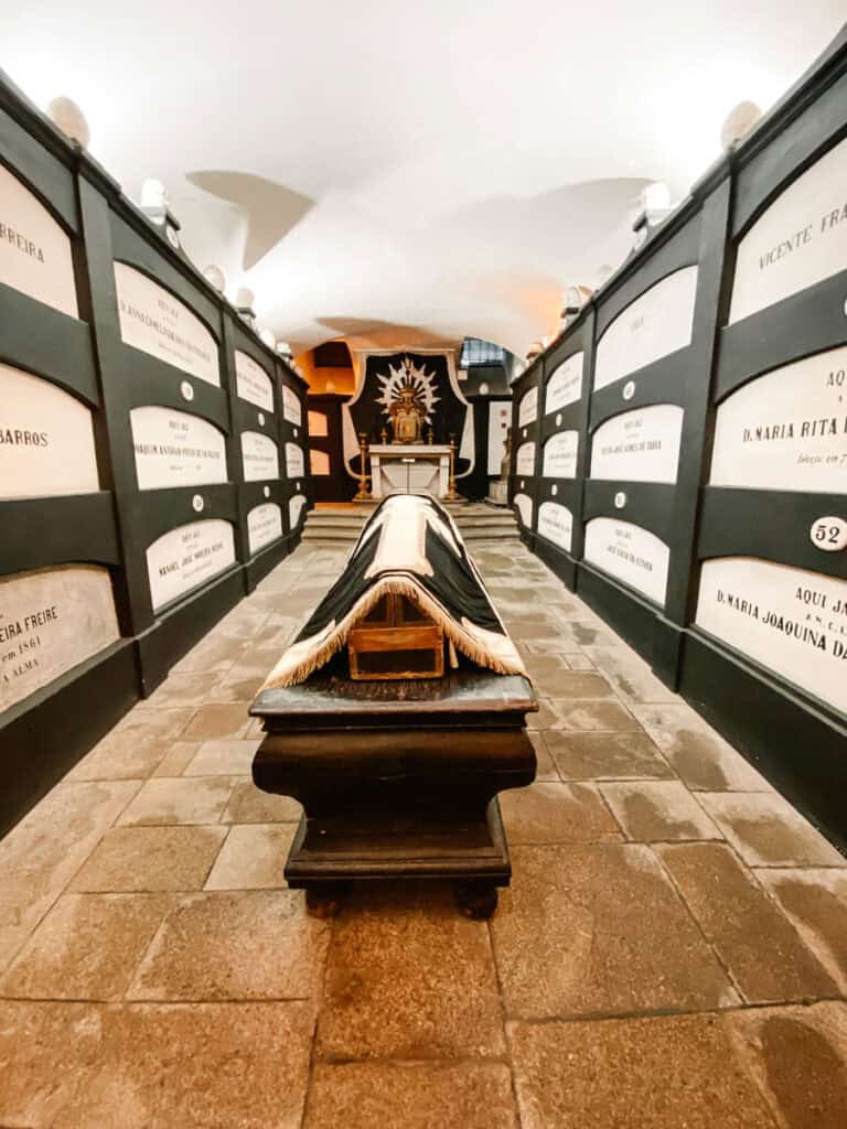 Cripta da igreja de São Francisco Portugal com corredor ladeado por nichos funerários identificados nas paredes e um caixão coberto ao centro. Ao fundo, um pequeno altar iluminado reforça a atmosfera silenciosa e histórica do espaço subterrâneo.