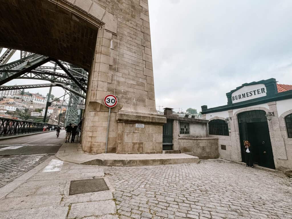 The Burmester Port Wine House stands beside the stone and iron bridge with "BURMESTER" displayed above its dark green entrance. A cobbled lane curves past the building while cyclists and pedestrians cross beneath the bridge arch.