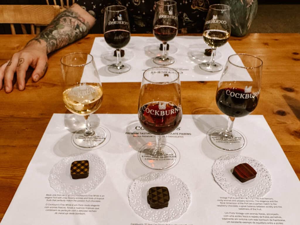 A Cockburn’s Port wine tasting with three glasses of pale gold amber and dark red port paired with individual chocolates on a wooden table. The glasses read "COCKBURN’S" and the tasting mat reads "COCKBURN’S PREMIUM TASTING WITH CHOCOLATE PAIRING".
