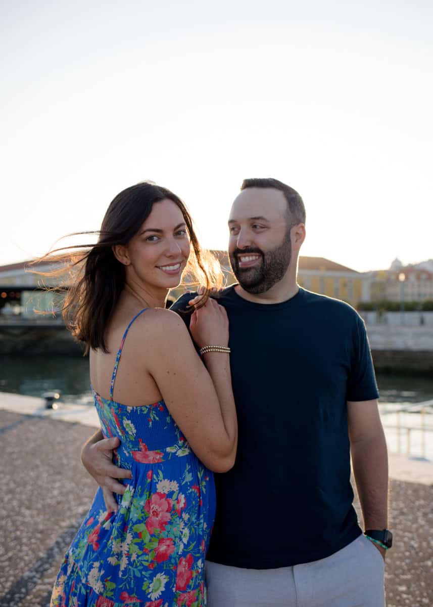 Stel omhelst elkaar aan het water in Lissabon terwijl de avondzon zacht licht door het haar van de vrouw laat schijnen. Tijdens deze fotoshoot voor stelletjes in Lissabon lachen ze ontspannen naar elkaar met een romantische sfeer langs de rivier op de achtergrond.