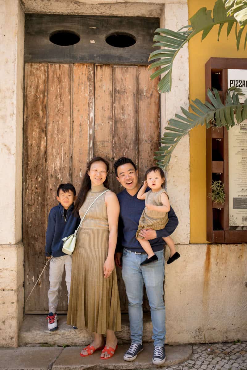 Gezin met twee jonge kinderen poseert lachend voor een rustieke houten deur in een zonnige straat in Lissabon tijdens een familie fotoshoot. De ouders dragen neutrale kleding terwijl tropische planten en warme gele muren zorgen voor een mediterrane sfeer.