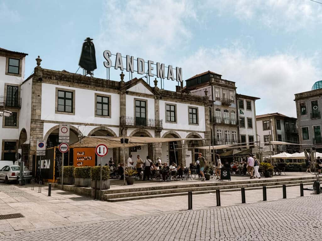 The Sandeman Port Wine House overlooks a lively cobbled square with outdoor café tables and historic buildings. Large rooftop letters read "SANDEMAN" beside the brand’s black caped figure.