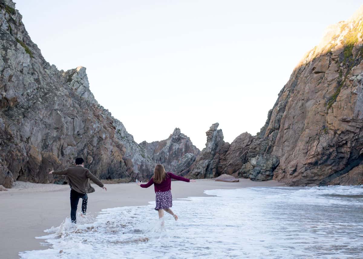 Casal corre descalço na beira do mar cercado por falésias rochosas na Praia da Ursa em Portugal. A paisagem selvagem e o clima descontraído criam um cenário romântico para um pedido de casamento na Praia da Ursa.