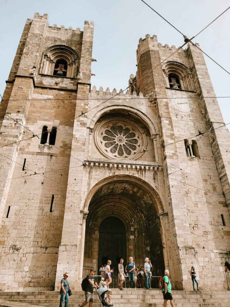 Fachada da Sé de Lisboa vista de baixo com duas torres de pedra e uma grande rosácea sobre a entrada principal. Visitantes estão reunidos nos degraus enquanto cabos de elétrico cruzam o céu em frente à catedral.