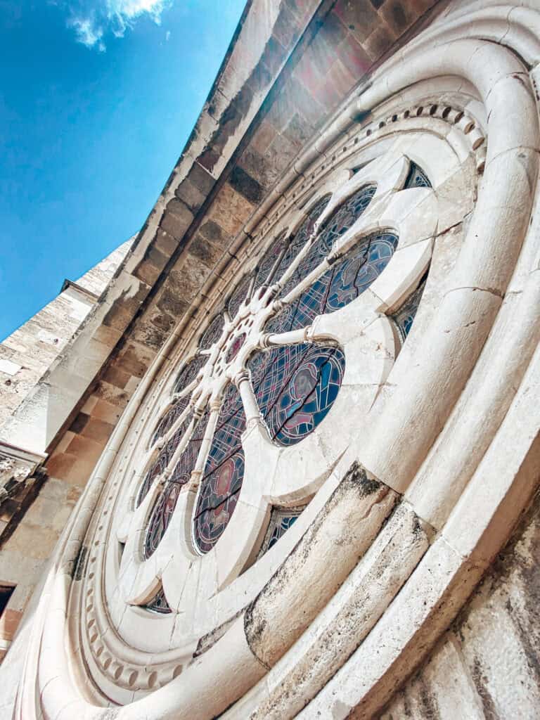 Detalhe da grande rosácea da Sé de Lisboa vista de baixo junto à fachada de pedra. O vitral circular exibe desenhos geométricos e figuras religiosas sob um céu azul intenso.