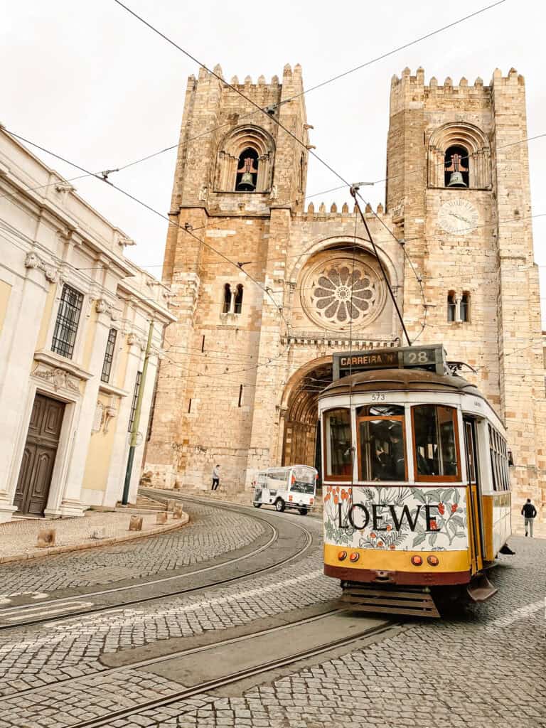 Elétrico 28 passa em frente à Catedral de Lisboa seguindo pelos carris de uma rua de calçada portuguesa. A fachada de pedra da Sé com as duas torres e a rosácea aparece ao fundo mostrando uma das formas mais tradicionais de como chegar à Catedral de Lisboa. No elétrico aparecem os textos "CARREIRA N. 28" "573" e "LOEWE".