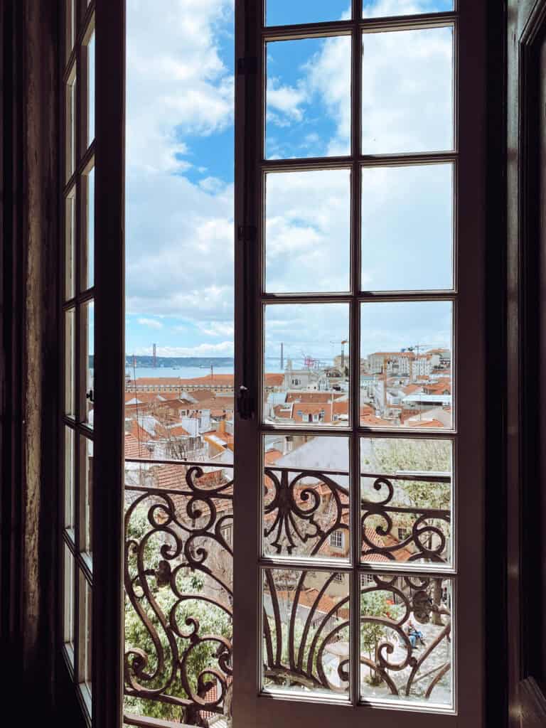 Vista da Catedral de Lisboa através de uma janela alta com grades de ferro decorativas. Telhados de terracota se espalham pela cidade com o rio Tejo e a Ponte 25 de Abril ao fundo sob céu azul com nuvens.