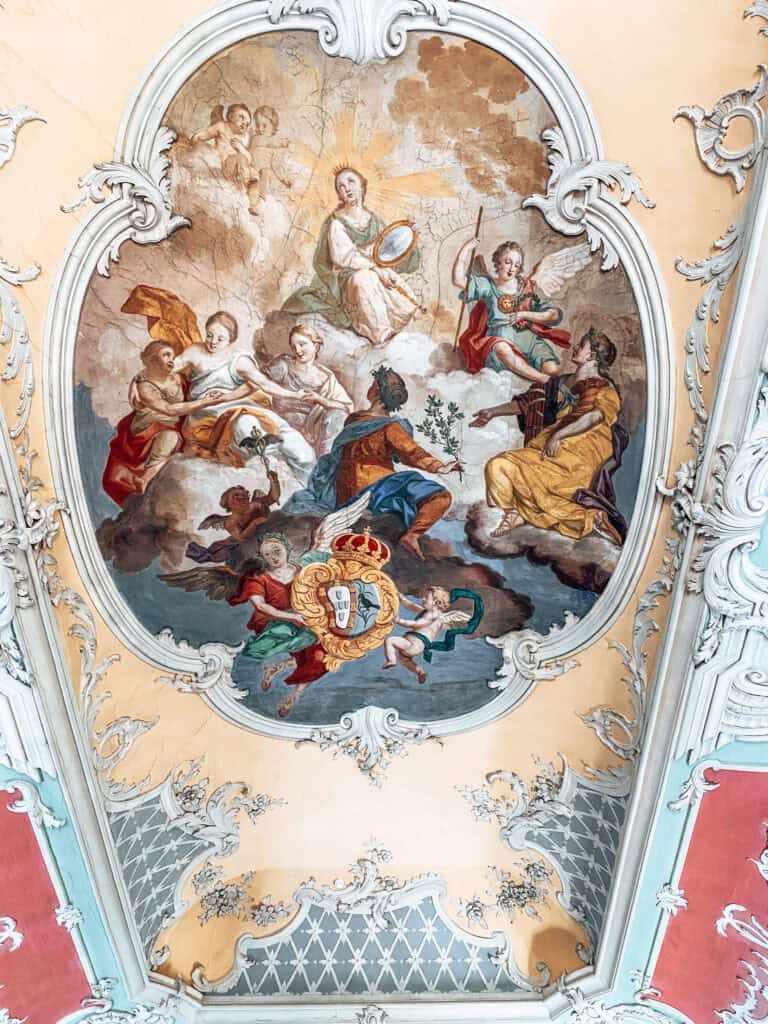 Teto ornamentado do tesouro da Catedral de Lisboa com pintura religiosa que reúne figuras celestiais e anjos entre nuvens. Um brasão coroado aparece na parte inferior da composição cercado por molduras decorativas em tons claros.