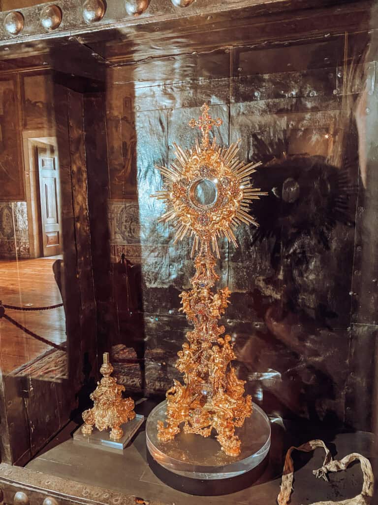 Peça litúrgica dourada ricamente ornamentada com cruz e estrutura em forma de raios exposta numa vitrine do acervo da Sé. O objeto religioso ajuda a ilustrar a história da Catedral de Lisboa e o seu património sacro preservado ao longo dos séculos.