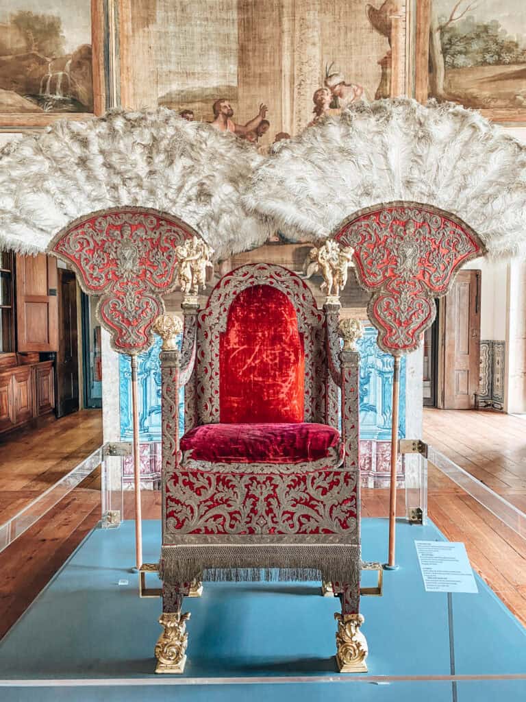 Trono cerimonial revestido de veludo vermelho e tecido bordado em prata acompanhado por dois grandes leques de penas brancas com detalhes dourados. A peça integra o património religioso ligado à história da Sé de Lisboa e está exposta numa sala decorada com pinturas e azulejos.