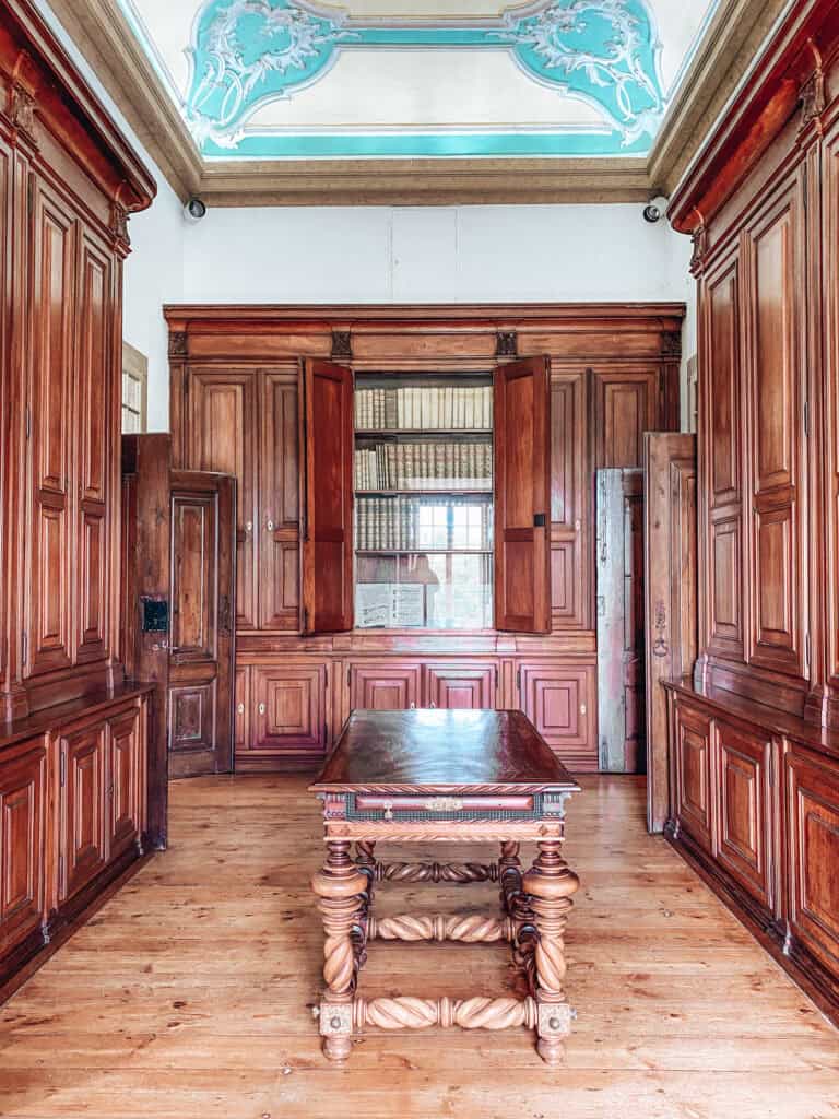 Sala do Tesouro da Sé de Lisboa com armários de madeira repletos de livros e uma mesa ornamentada ao centro. O teto decorado em azul turquesa e branco contrasta com a madeira escura e reforça o caráter histórico do espaço.