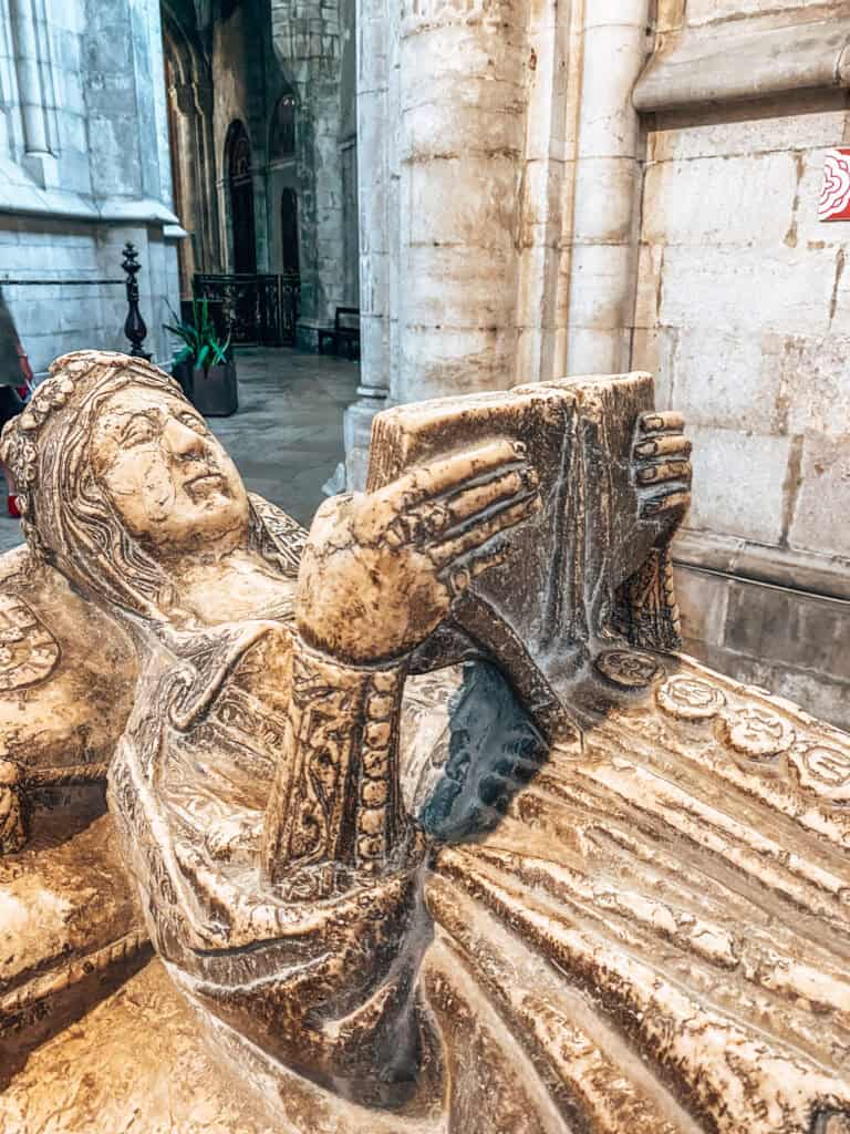 Túmulo medieval em pedra com figura reclinada segurando um livro aberto diante do peito e vestes detalhadamente esculpidas. A sepultura preservada no interior da catedral integra os monumentos funerários ligados à história da Sé de Lisboa.