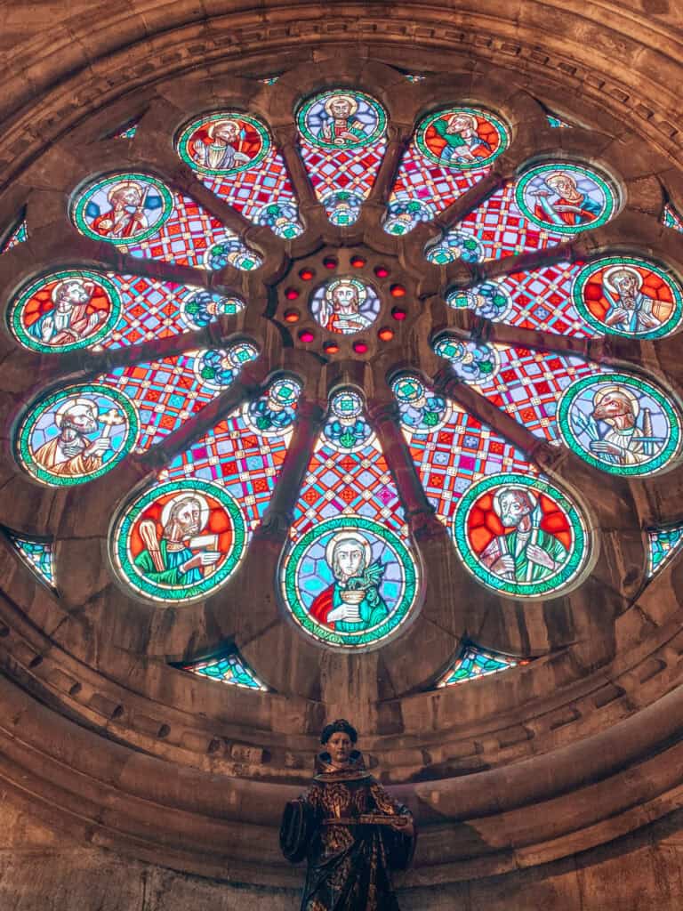 Rosácea colorida no interior da Catedral de Lisboa com vitrais que representam figuras religiosas em tons de vermelho azul e verde. Uma estátua religiosa aparece abaixo do vitral destacando a riqueza artística e espiritual do espaço.