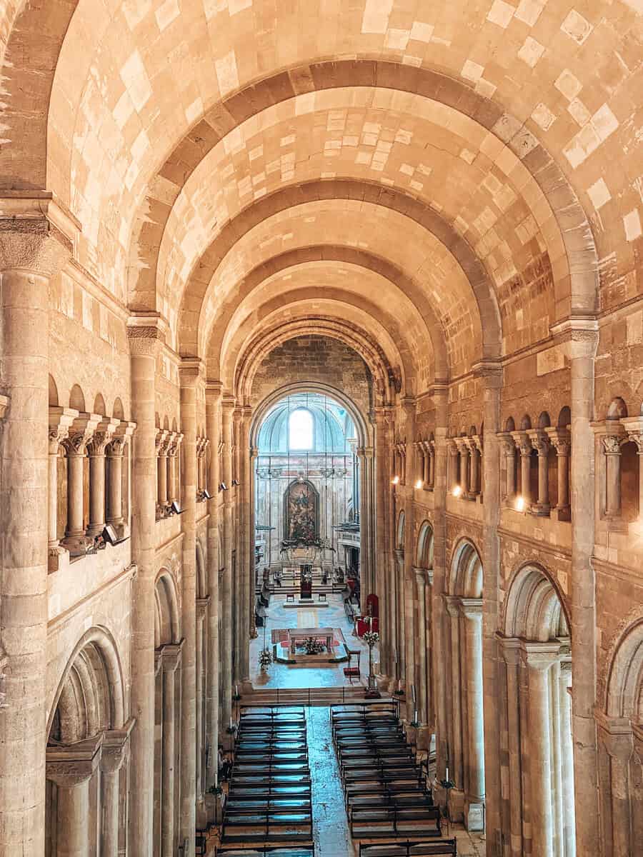 Interior da Catedral de Lisboa visto de um nível elevado com a nave central e os bancos conduzindo até ao altar. Grandes arcos de pedra e colunas românicas destacam a arquitetura histórica do interior da Sé.