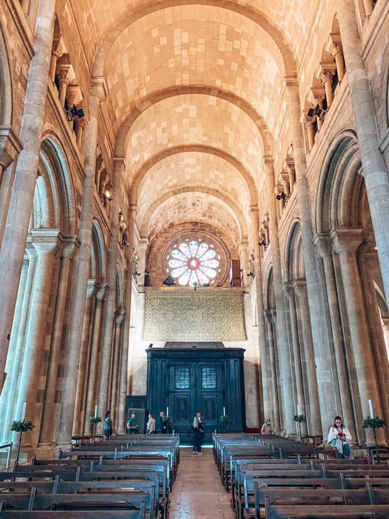 Nave da Sé de Lisboa vista entre fileiras de bancos de madeira sob uma alta abóbada de pedra sustentada por arcos e colunas românicas. Ao fundo a rosácea colorida ilumina o interior acima de uma grande estrutura de madeira escura.