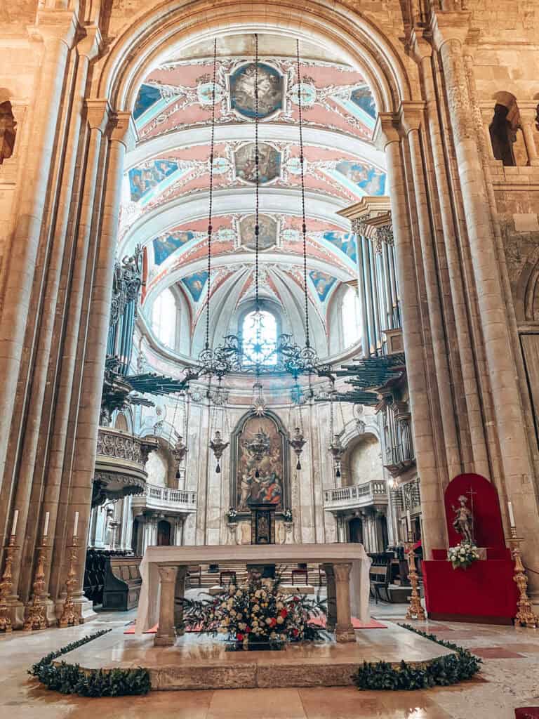 Interior de uma capela da Sé de Lisboa com altar de pedra decorado com flores e grandes castiçais dourados. Ao fundo aparecem uma pintura religiosa e um teto abobadado ornamentado em tons de rosa azul e branco.