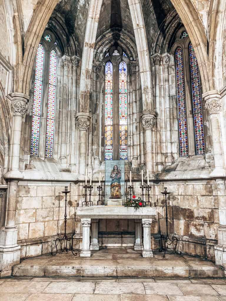 Capela na Catedral de Lisboa com altar de pedra ao centro e uma pequena imagem religiosa protegida por vidro. Altos arcos góticos e vitrais estreitos e coloridos iluminam o espaço histórico.