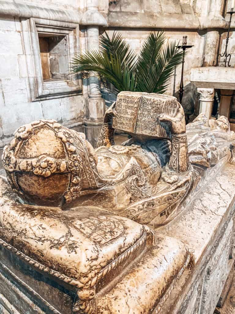 Túmulo esculpido em pedra com a figura de um religioso usando mitra e segurando um livro aberto sobre o peito. A peça funerária preservada no interior da Sé ajuda a contar a história da Catedral de Lisboa por meio dos seus monumentos e sepulturas medievais.