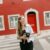 Lisbon photographer standing on a zebra crossing and holding a camera in front of a red building with white trimmed windows and a matching red door. She looks off to the side in a relaxed portrait that introduces the photographer behind the service.