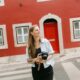 Lisbon photographer standing on a zebra crossing and holding a camera in front of a red building with white trimmed windows and a matching red door. She looks off to the side in a relaxed portrait that introduces the photographer behind the service.