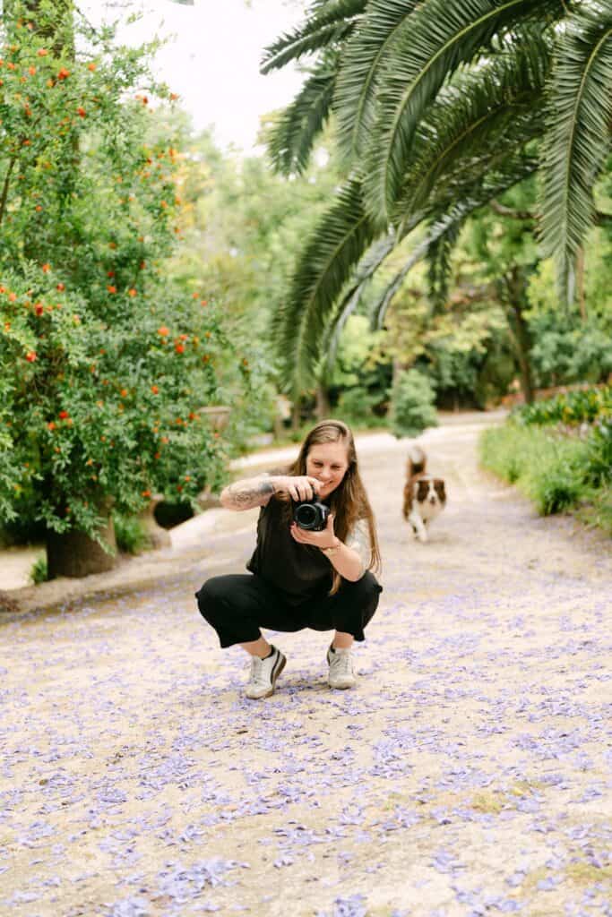 Lisbon photographer crouching on a garden path covered with purple flower petals while taking a photo with her camera. Palm leaves, flowering trees, and a dog walking in the background create a relaxed outdoor photoshoot setting.