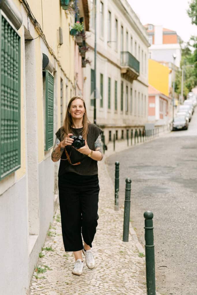 Photographer in Lisbon walking along a quiet cobbled street while holding a camera and smiling at the camera. Colourful buildings, green shutters, and a steep Lisbon road frame the professional photoshoot setting.