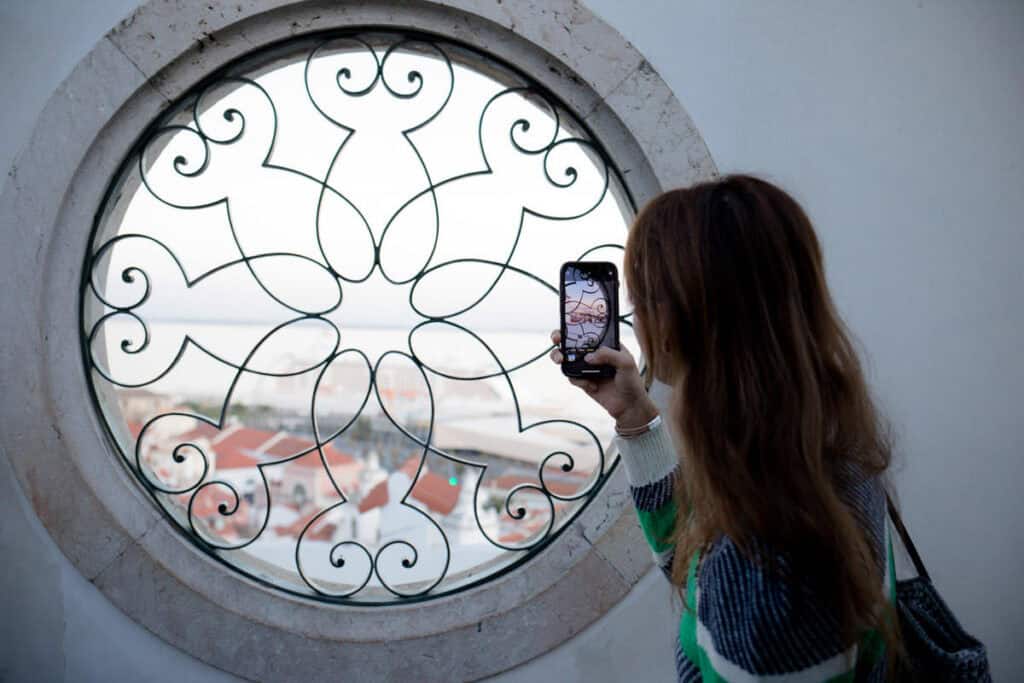 A woman photographs an ornate circular iron window that frames red tiled rooftops and the Tagus River beyond. The scene demonstrates natural framing during Photography Workshops in Lisbon.