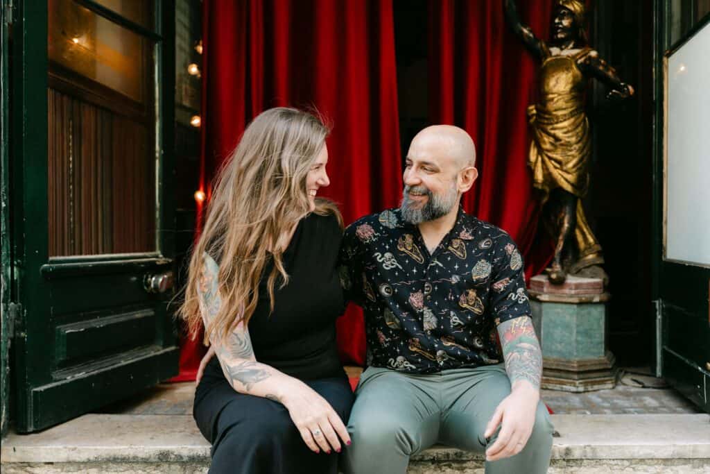 Marga en Ulisses zitten lachend samen voor de ingang van een sfeervol gebouw in Lissabon.