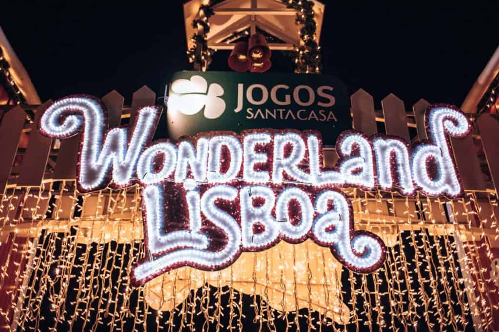Letreiro luminoso do Wonderland Lisboa à noite, decorado com luzes de Natal brancas e douradas. Por cima, o logótipo dos Jogos Santa Casa e sinos natalícios decoram a entrada do evento.