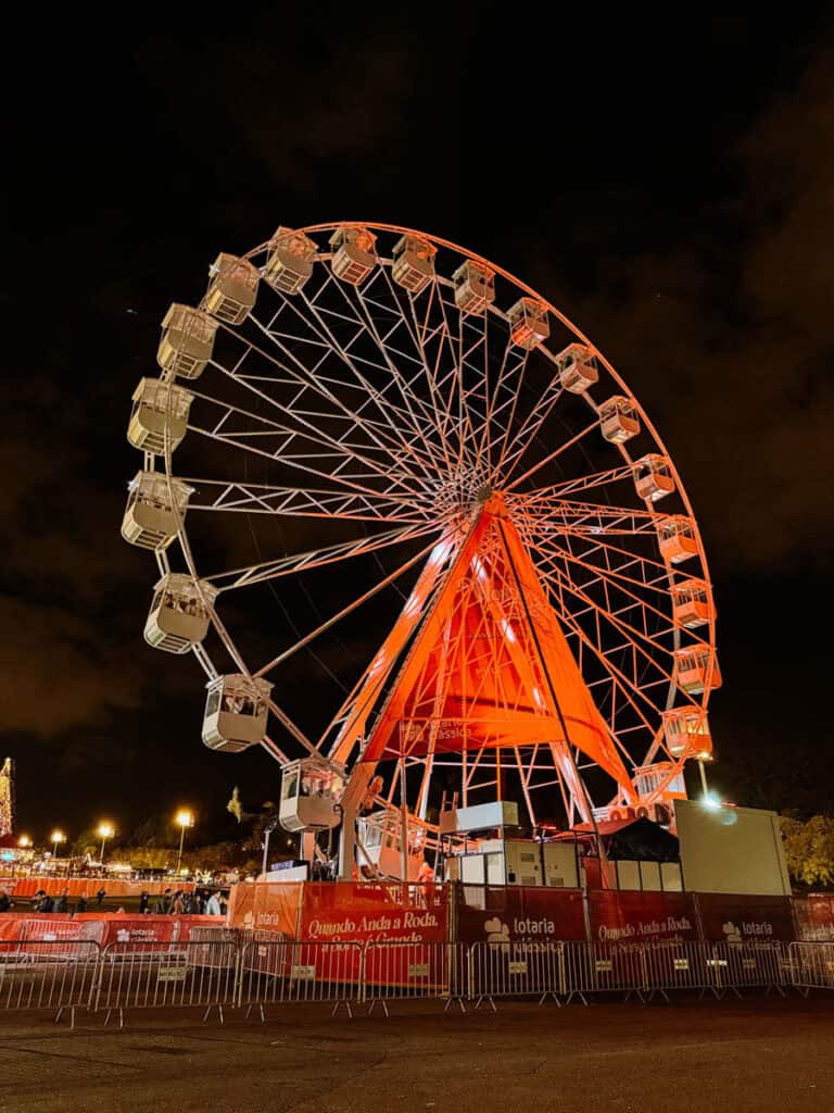 Roda gigante do Wonderland Lisboa iluminada em tons de laranja durante a noite, com várias cabines suspensas e uma estrutura metálica imponente. A atração destaca-se no mercado de Natal em Lisboa, rodeada por luzes natalícias e visitantes.