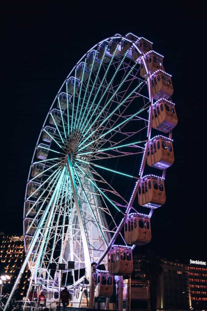 Roda gigante do Wonderland Lisboa iluminada em tons de azul turquesa e roxo durante a noite, com cabines fechadas e visitantes a bordo. A estrutura destaca-se contra o céu escuro, com os edifícios da Praça Marquês de Pombal iluminados ao fundo.