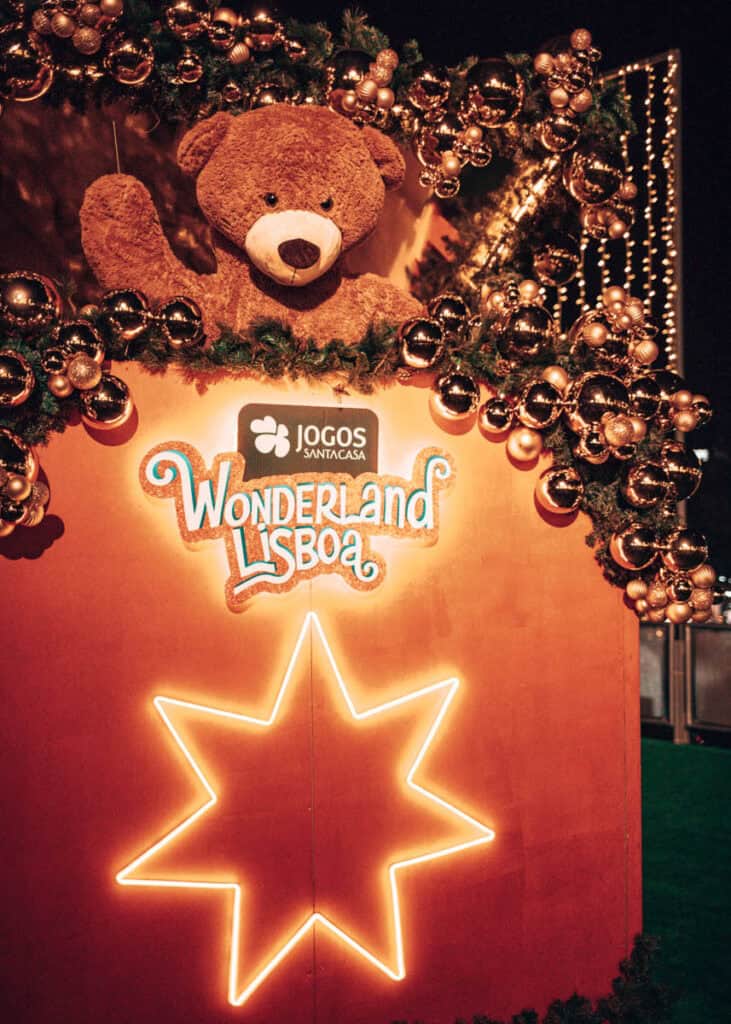 Urso de peluche castanho gigante rodeado de grinaldas verdes e bolas de Natal douradas no Wonderland Lisboa. Por baixo, o logótipo luminoso do evento e dos Jogos Santa Casa destaca-se sobre um painel cor de rosa com uma grande estrela iluminada.