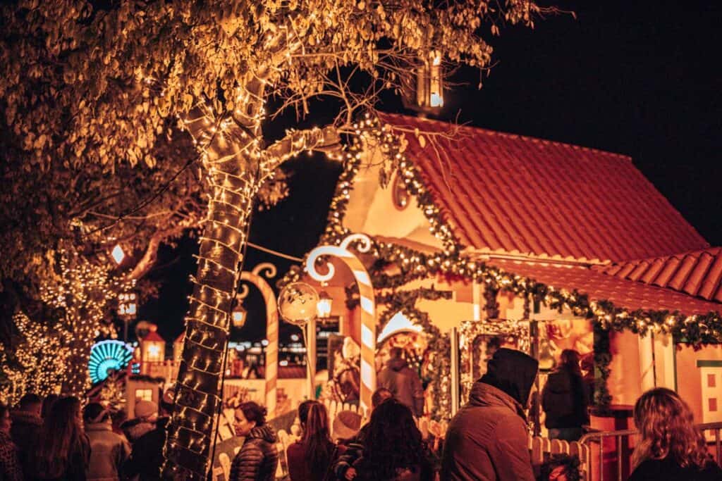 Visitantes passeiam pelo Wonderland Lisboa à noite, junto a uma casinha de Natal com telhado vermelho, grinaldas verdes e luzes douradas. Árvores iluminadas e decorações em forma de bengalas doces criam um ambiente natalício acolhedor.