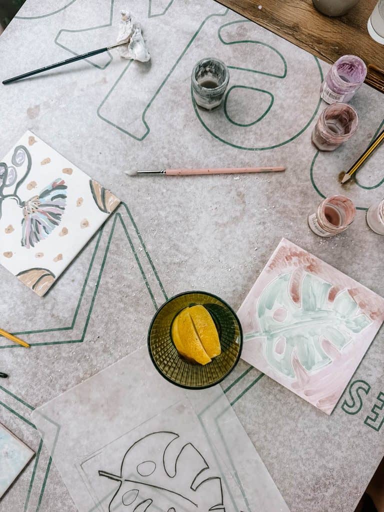 An overhead view of an Azulejo workshop in Lisbon with painted ceramic tiles featuring a colourful bird and a pale green leaf. Pots of paint brushes tracing paper and a bowl with a cut lemon are scattered across the worktable.