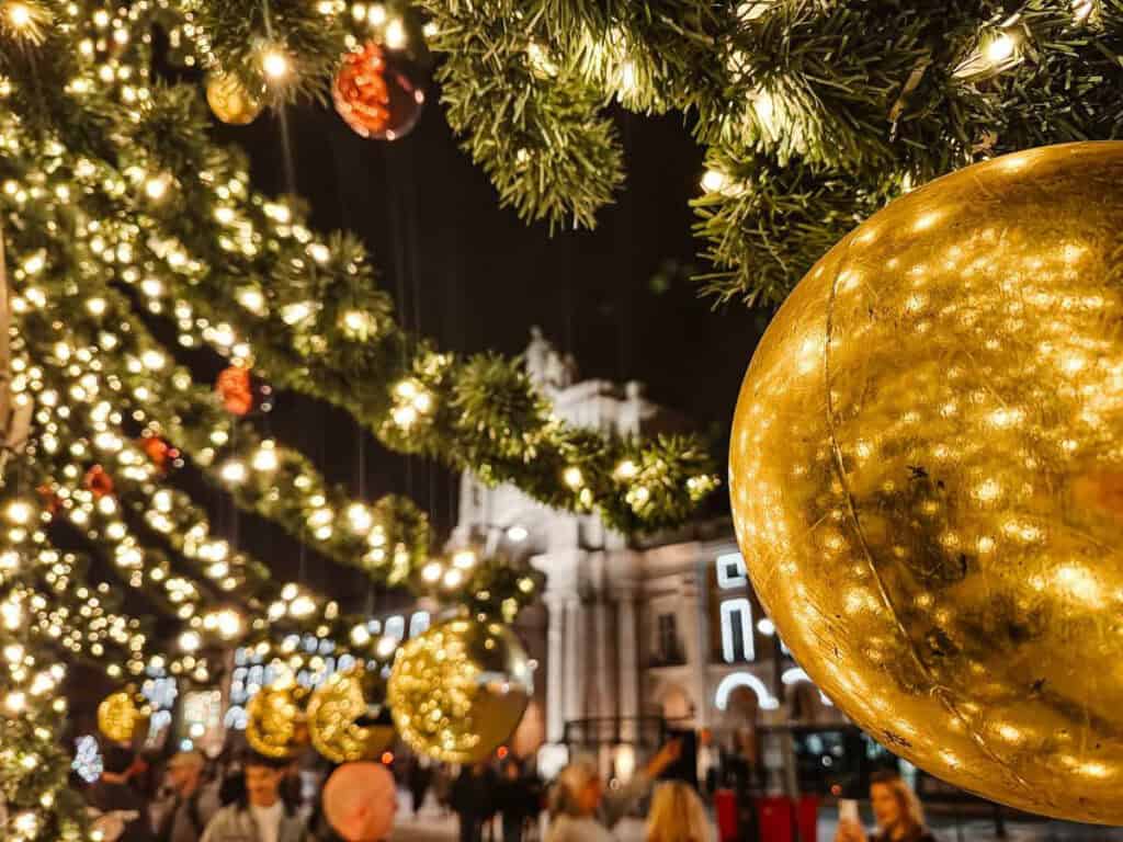Grinaldas verdes iluminadas com luzes douradas e grandes bolas de Natal suspensas junto ao Arco da Rua Augusta em Lisboa. A decoração festiva na Praça do Comércio é um dos locais onde ver as luzes de Natal em Lisboa.