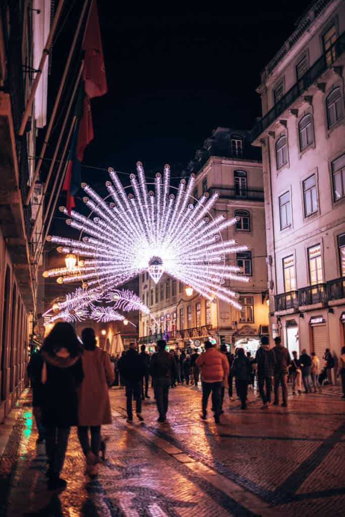 Rua pedonal da Baixa de Lisboa decorada com luzes de Natal brancas e lilases em forma de leques luminosos suspensos entre os edifícios. Várias pessoas passeiam à noite sob a iluminação de Natal em Lisboa que se reflete na calçada portuguesa.