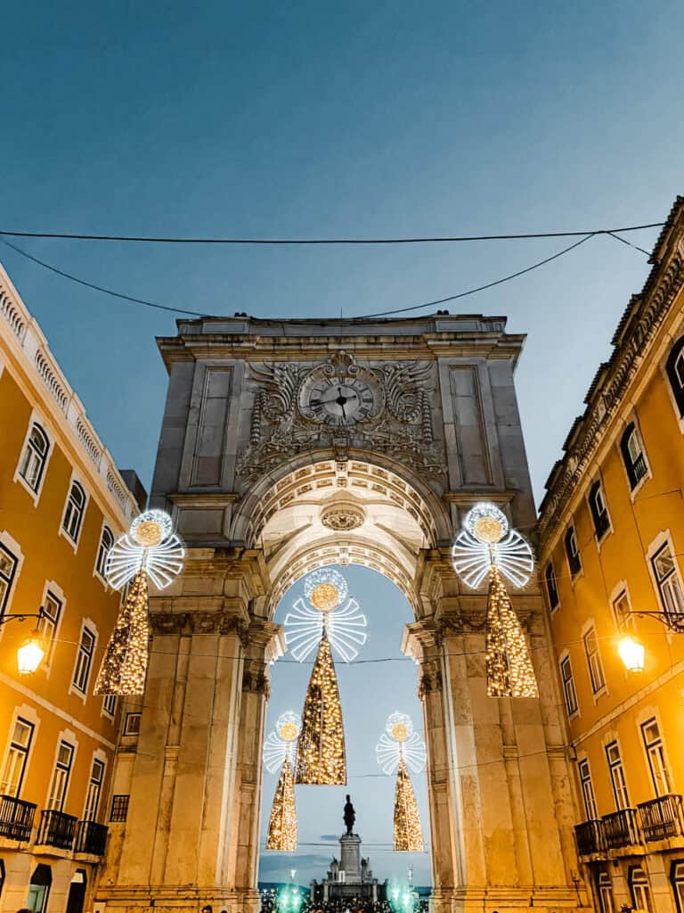 Arco da Rua Augusta em Lisboa ao anoitecer com decorações de Natal suspensas em forma de anjos iluminados com luzes brancas e douradas. Ao fundo avista-se a estátua de D. José I na Praça do Comércio, um dos locais onde ver as luzes de Natal em Lisboa.